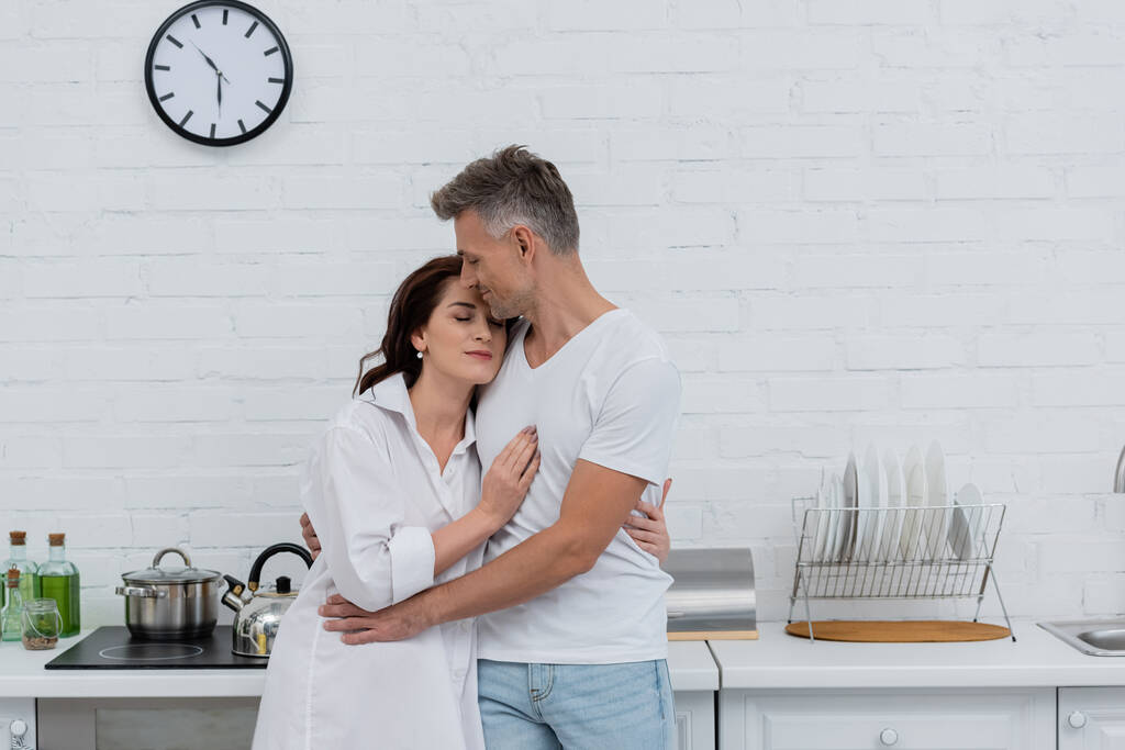 Homem com olhos fechados abraçando a esposa morena na camisa na cozinha  - Foto, Imagem