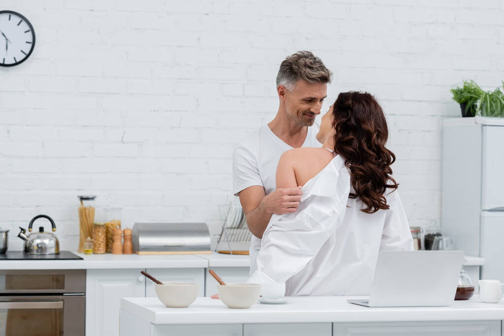 Позитивний чоловік знімає сорочку від брюнетки дружини біля ноутбука і кави на кухні
  - Фото, зображення