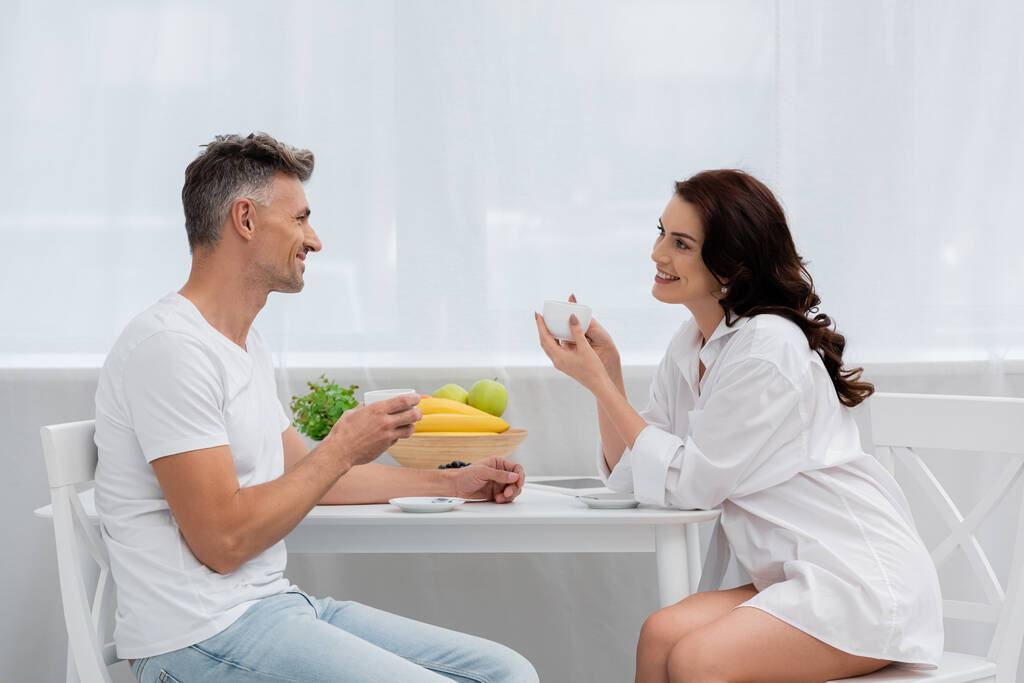 Улыбающаяся женщина в рубашке держит чашку кофе рядом с мужем и фрукты на кухне  - Фото, изображение