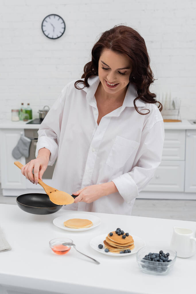Mulher sorridente na camisa cozinhar panquecas perto de mirtilos na cozinha  - Foto, Imagem