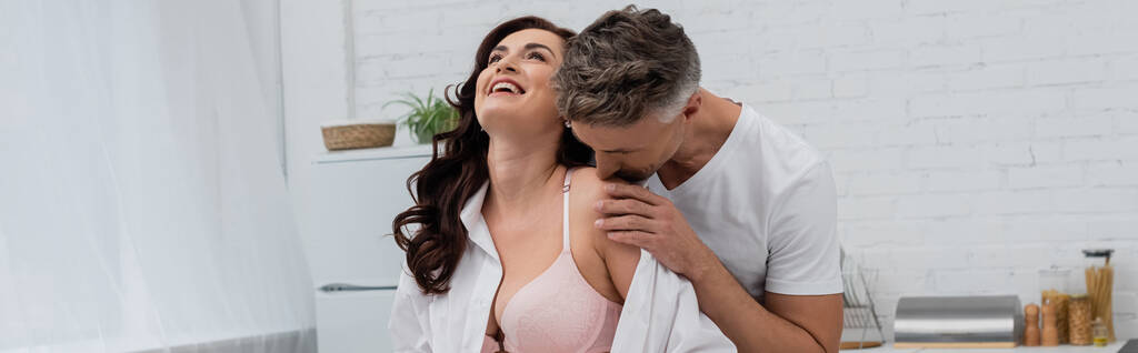 Homme embrassant épaule de femme souriante en soutien-gorge et chemise dans la cuisine, bannière  - Photo, image