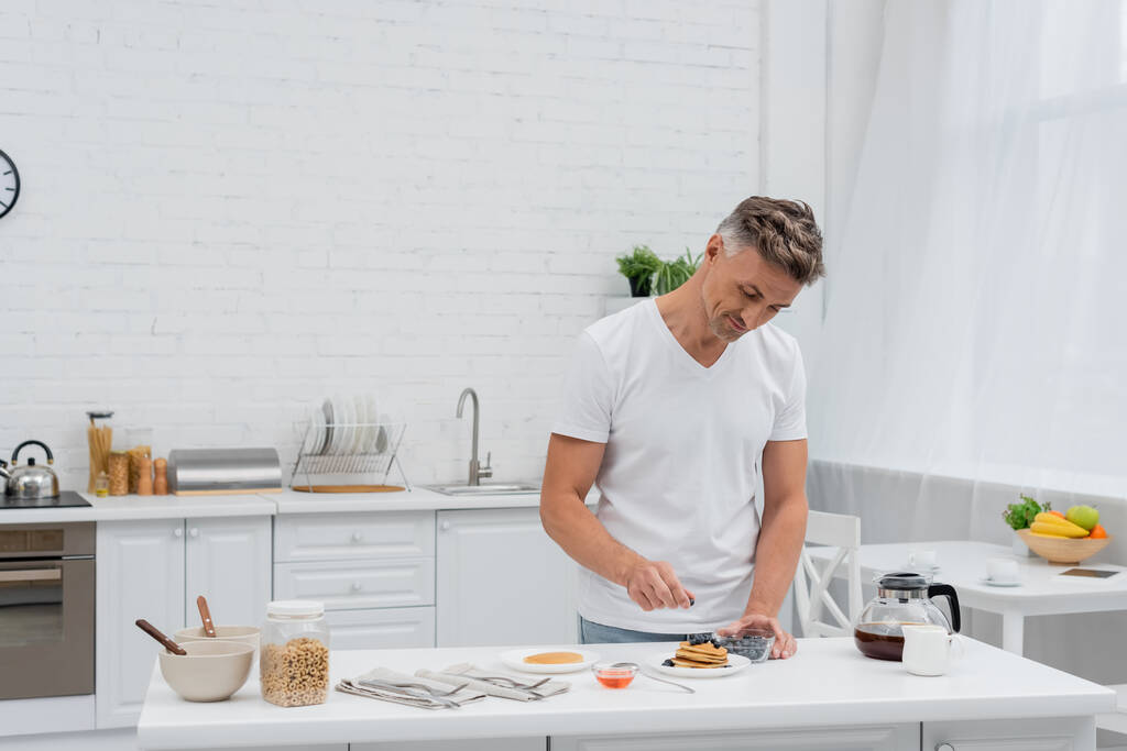 Homem segurando mirtilos perto de deliciosas panquecas e café na cozinha  - Foto, Imagem