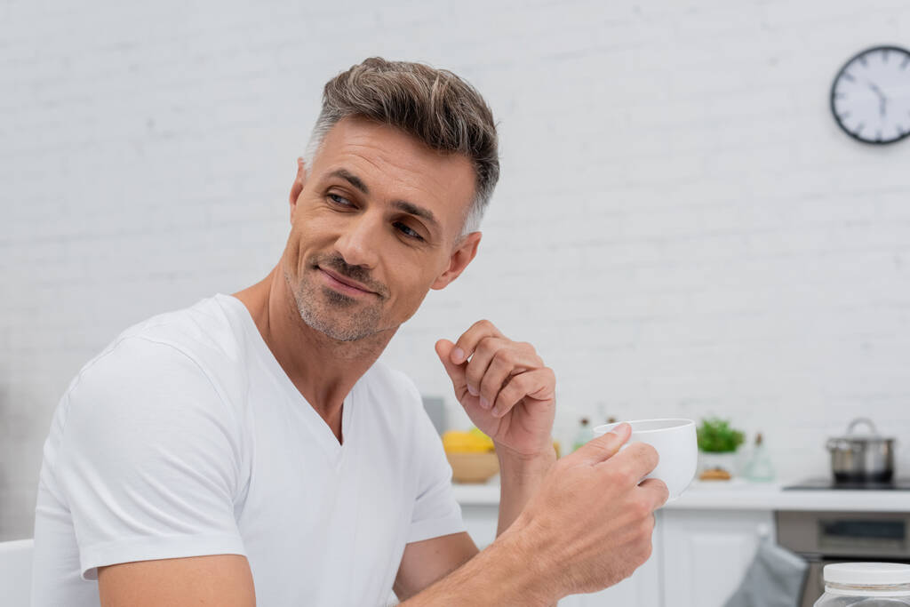 Мужчина в футболке держит чашку кофе на кухне дома  - Фото, изображение