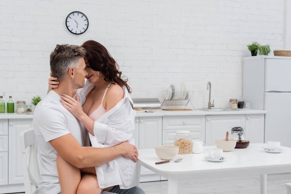 Sexy woman in bra and shirt kissing husband near breakfast in kitchen  - Photo, Image