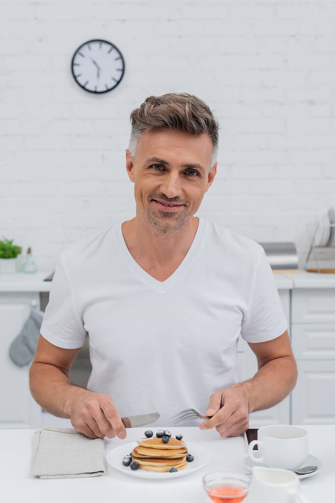 Усміхнений чоловік дивиться на камеру біля млинців з чорницею на кухні
  - Фото, зображення