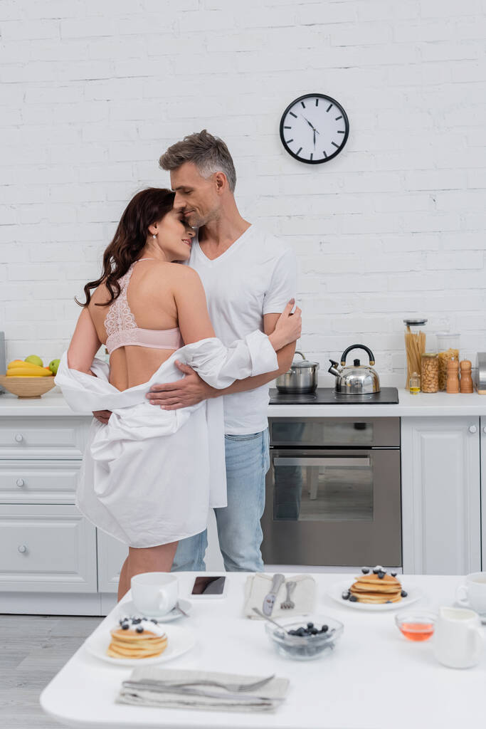 Side view of sexy woman in bra and shirt hugging husband near blurred breakfast at home  - Photo, Image
