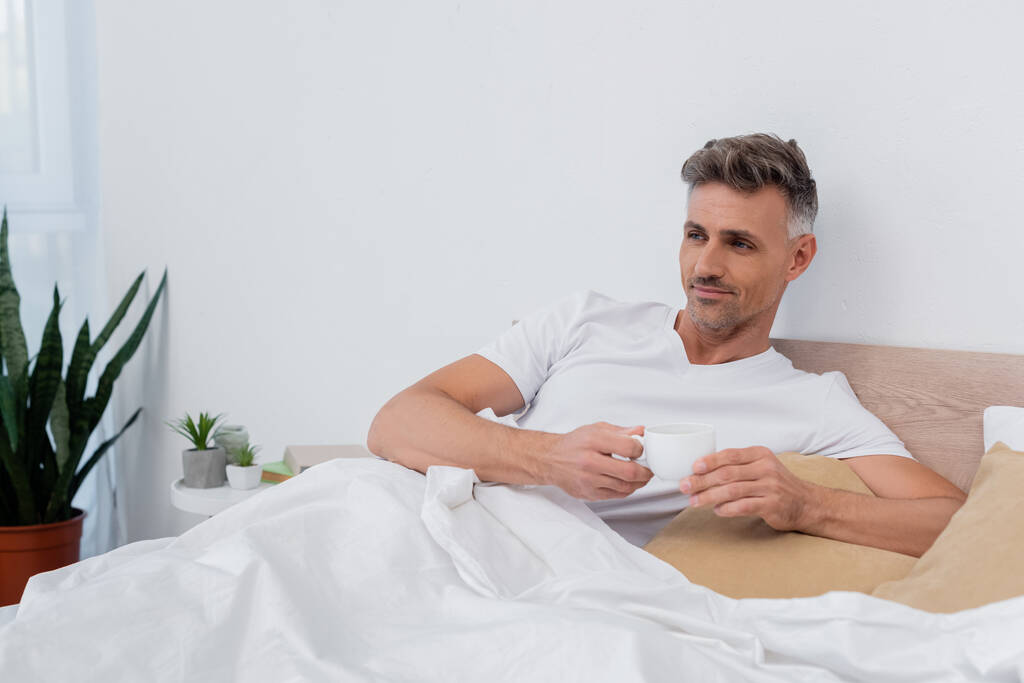 Мужчина в футболке, держа чашку кофе, лежа дома на кровати  - Фото, изображение