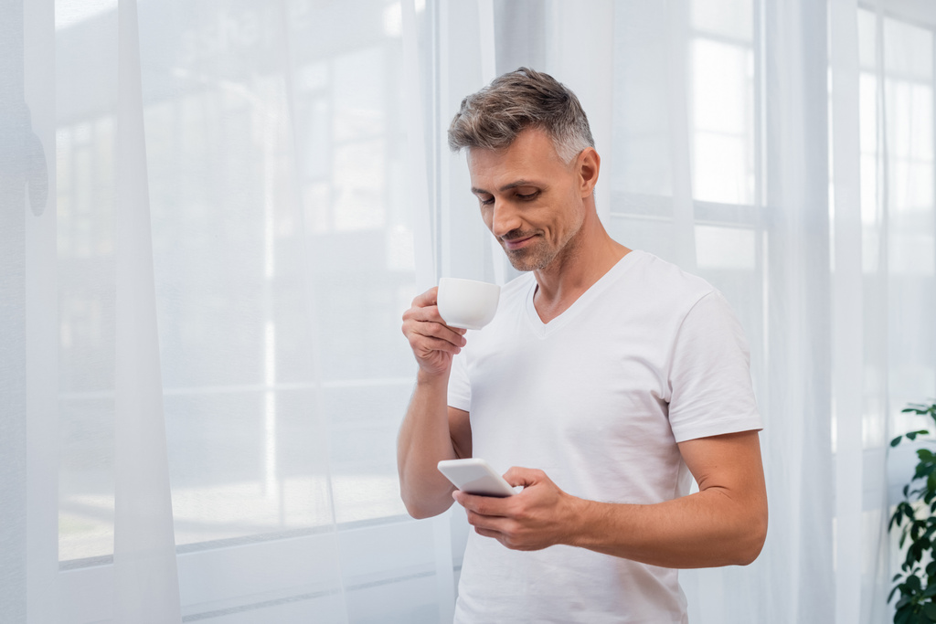 Mies valkoisessa t-paidassa kännykällä ja kahvikupin pitäminen kotona  - Valokuva, kuva