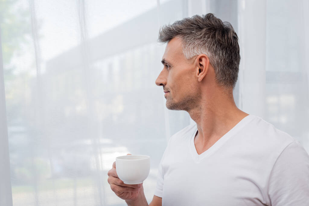 Vue latérale de l'homme en t-shirt blanc tenant tasse de café près des rideaux à la maison  - Photo, image