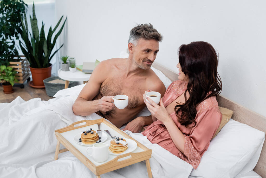 Sonriente hombre sin camisa sosteniendo taza de café cerca de esposa morena y panqueques en bandeja en el dormitorio  - Foto, imagen