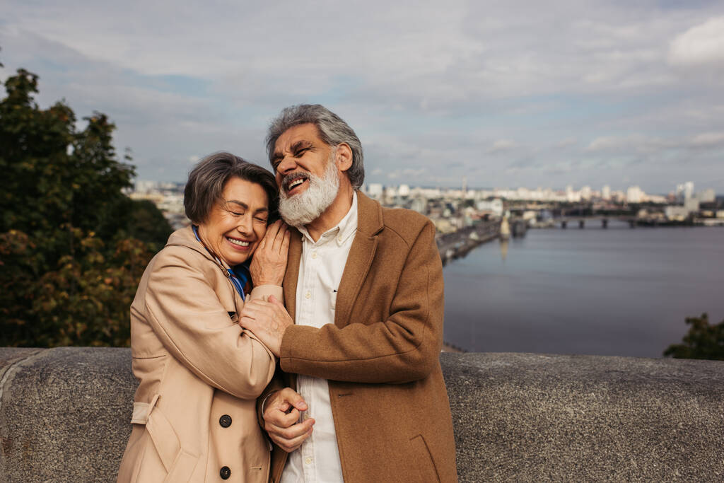senior vrouw leunend op schouder van man in jas en lachen in de buurt van rivier  - Foto, afbeelding