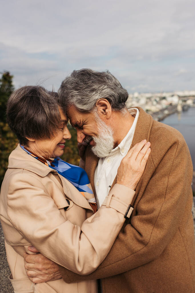 senior woman hugging with bearded husband in coat and smiling near river - Photo, Image