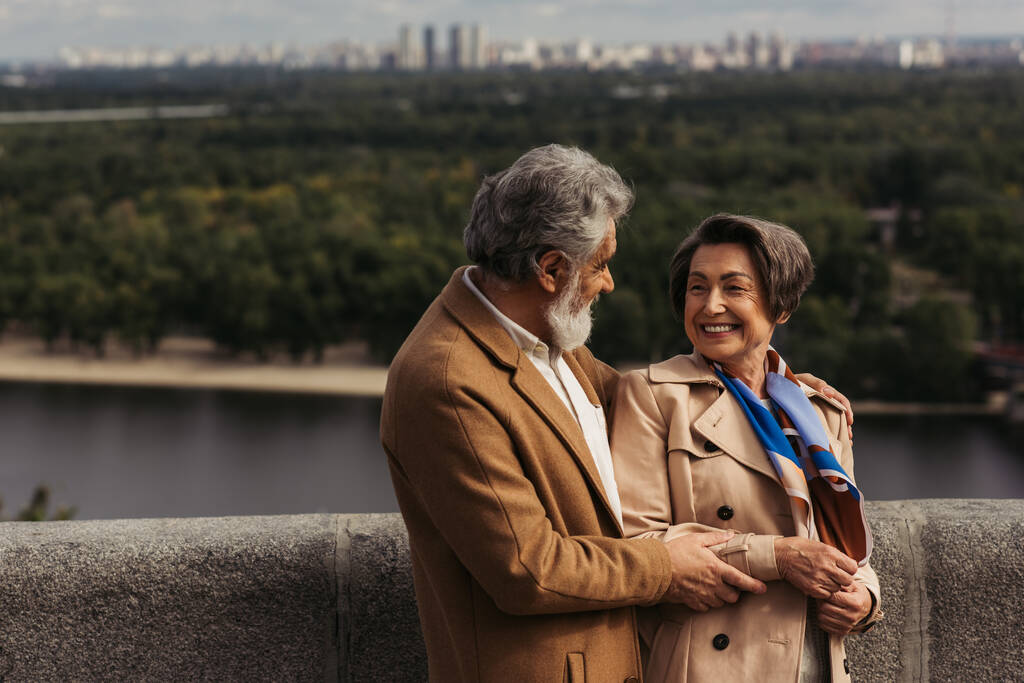 bärtiger älterer Mann lächelt, während er fröhliche Frau auf Brücke in der Nähe des Flusses umarmt - Foto, Bild