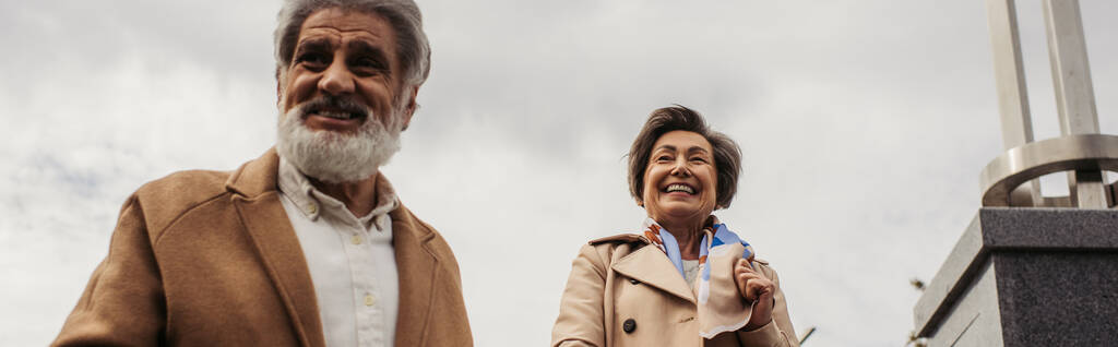 Mutlu karısıyla gülümseyen ceketli yaşlı adam bulutlu gökyüzüne ve afişe karşı - Fotoğraf, Görsel