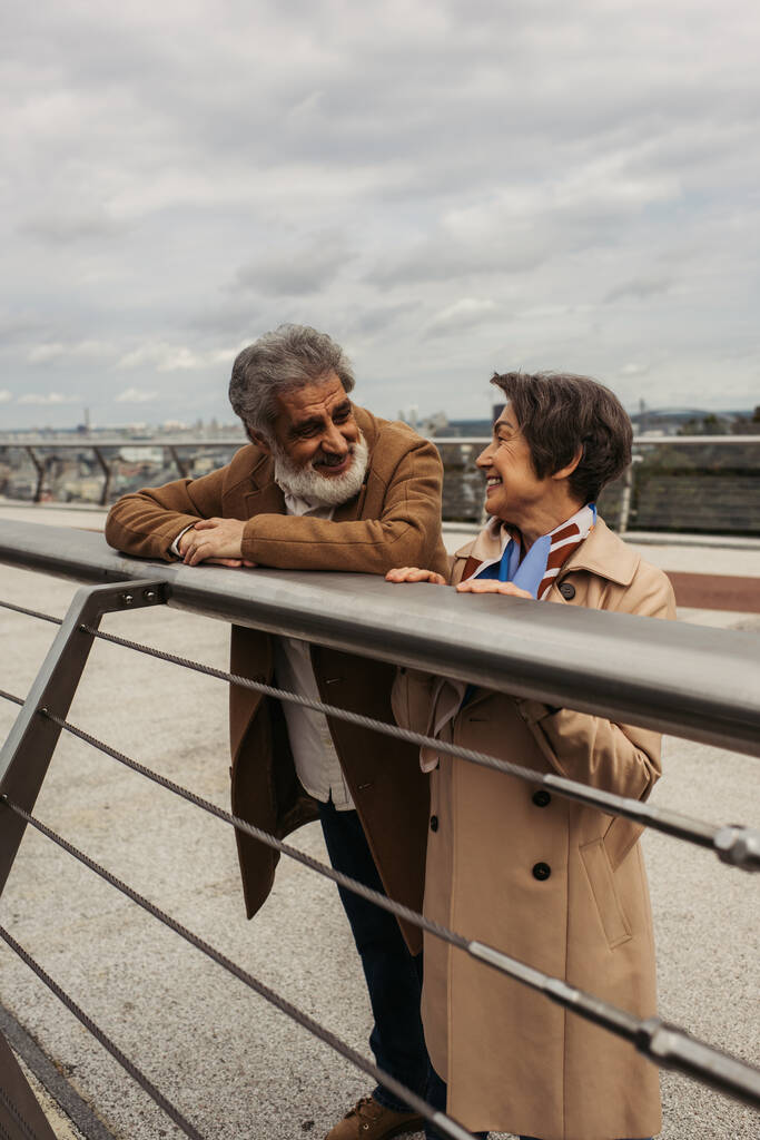 šťastný starší muž v kabátě při pohledu na potěšenou ženu, zatímco se opírá o zábradlí mostu  - Fotografie, Obrázek