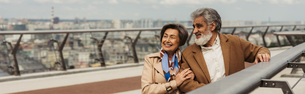 ευτυχισμένη ηλικιωμένη γυναίκα αγκαλιάζει γενειοφόρο και χαρούμενο σύζυγο κοντά στο κιγκλίδωμα γέφυρα, πανό - Φωτογραφία, εικόνα