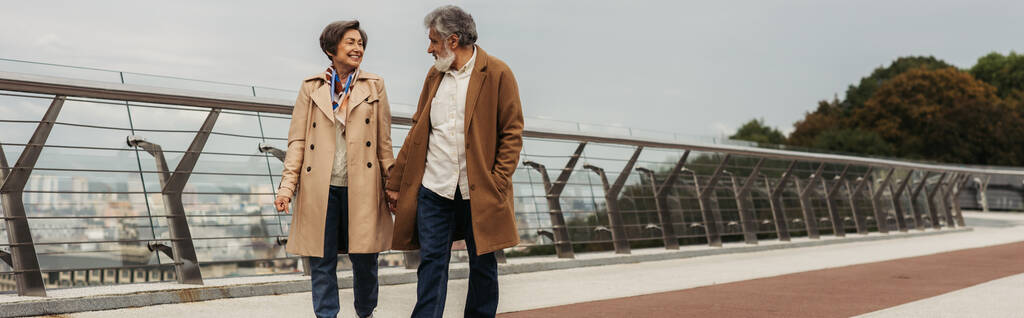 χαρούμενο ζευγάρι ηλικιωμένων σε μπεζ παλτά κρατώντας τα χέρια και περπατώντας στη γέφυρα κοντά στο κιγκλίδωμα, λάβαρο - Φωτογραφία, εικόνα