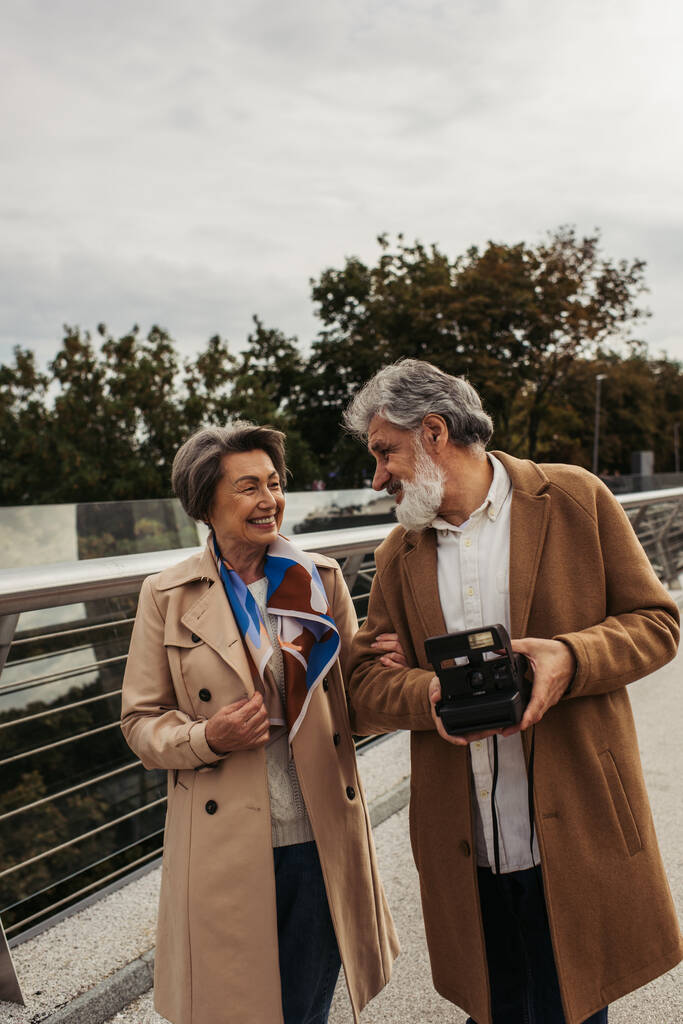 onnellinen vanhempi mies tilalla vintage kamera lähellä hymyilevä vaimo sillalla  - Valokuva, kuva
