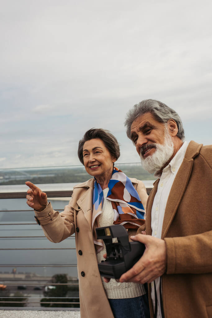 feliz hombre mayor sosteniendo la cámara vintage cerca de la esposa sonriendo y señalando con el dedo en el puente  - Foto, imagen