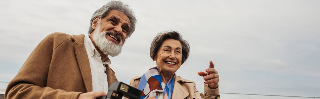 parrakas ja vanhempi mies, jolla on vintage-kamera lähellä vaimoa hymyillen osoittaen sormella, banneri - Valokuva, kuva