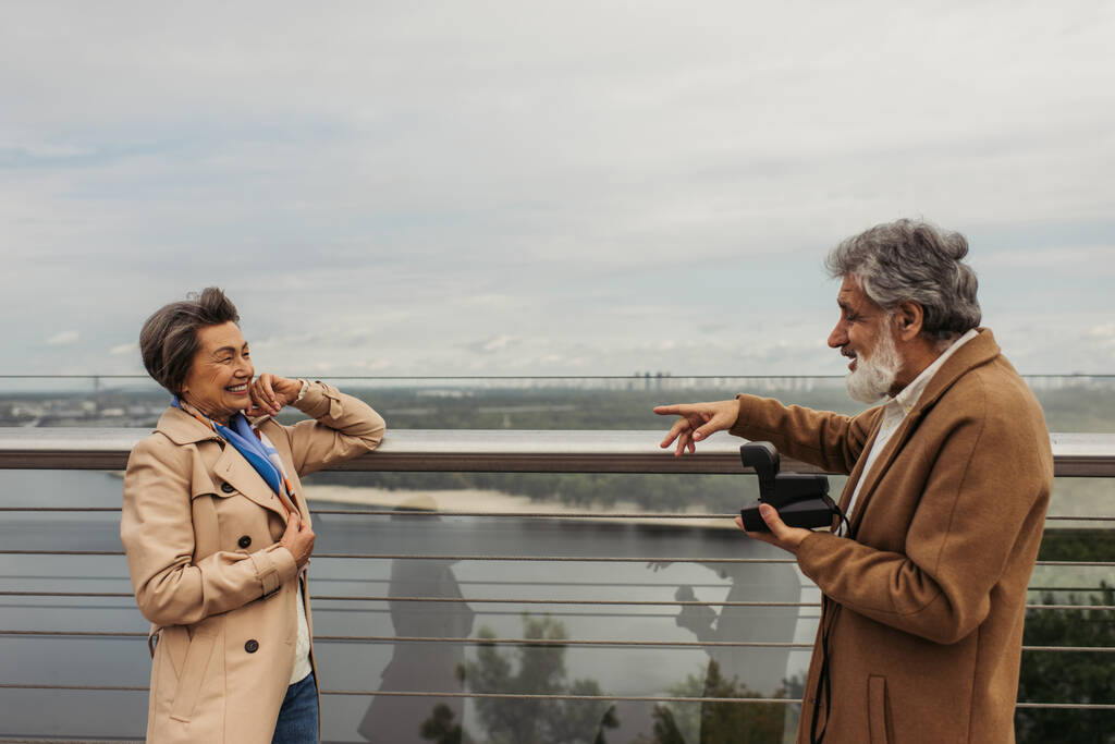 side view of bearded man holding vintage camera and gesturing near happy senior woman posing near guard rail  - Photo, Image