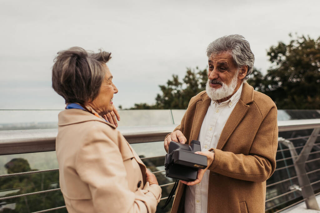 Senior bärtiger Mann hält Oldtimer-Kamera und spricht mit Frau, die in der Nähe der Brückenleitplanke steht  - Foto, Bild