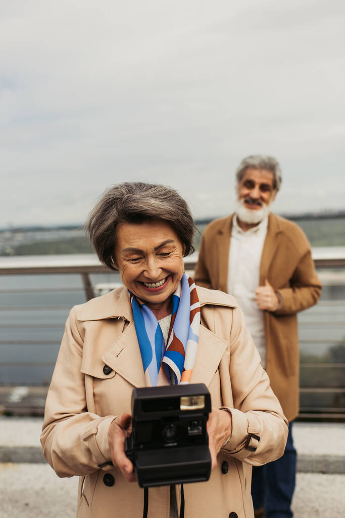 šťastný senior žena držení vintage kamera v blízkosti rozmazaný manžel na pozadí  - Fotografie, Obrázek