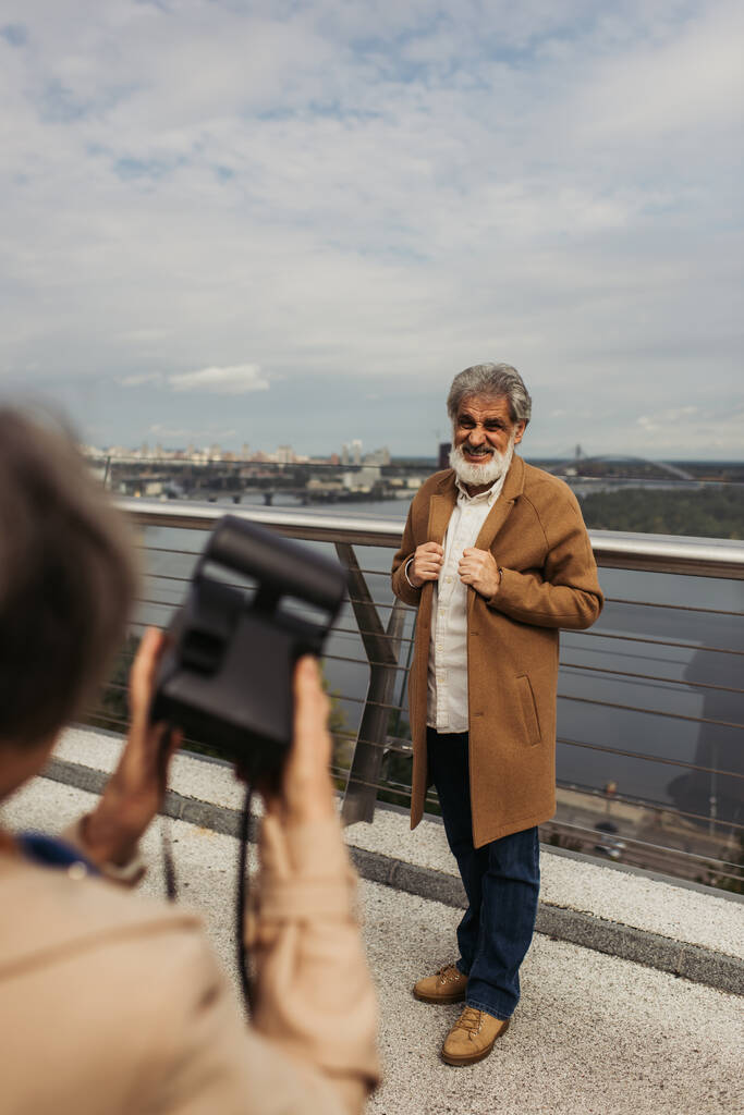 blurred woman holding vintage camera while taking photo of bearded senior husband on bridge near river - Photo, Image