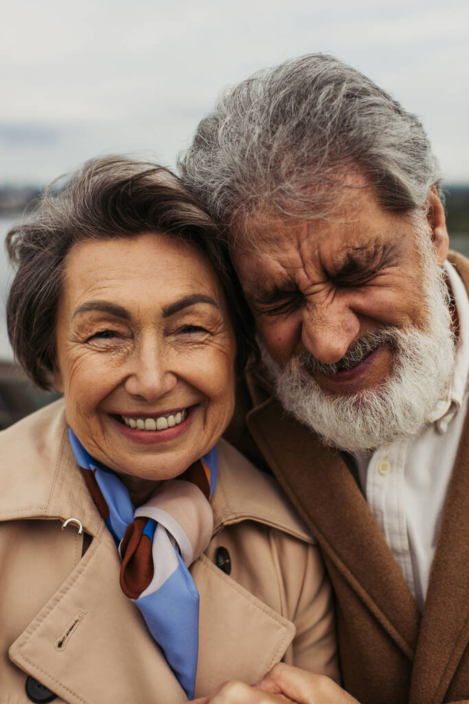 portrét šťastného staršího muže a pozitivní ženy v béžových kabátech usmívající se venku  - Fotografie, Obrázek