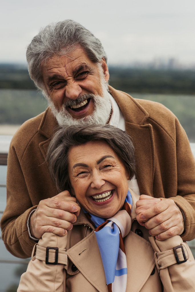 Porträt eines glücklichen älteren Mannes und einer fröhlichen Frau in beigen Mänteln, die lächelnd in die Kamera blicken  - Foto, Bild