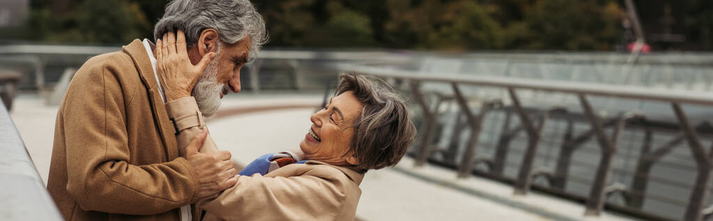 vrolijke oudere vrouw knuffelen bebaarde man in beige jas op brug in de buurt van guard rail, banner - Foto, afbeelding