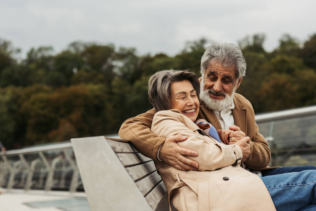 счастливый пожилой мужчина обнимает радостную жену, сидя на скамейке на открытом воздухе  - Фото, изображение