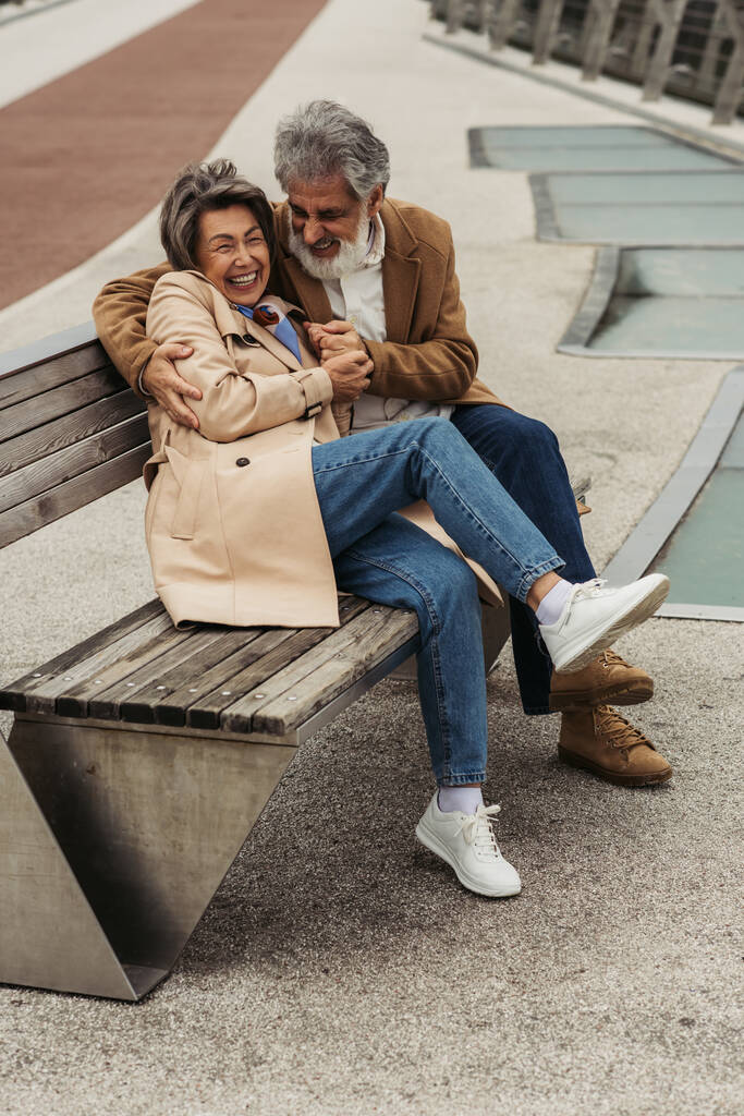 piena lunghezza di felice uomo anziano abbracciare allegra moglie ridendo mentre seduto sulla panchina - Foto, immagini