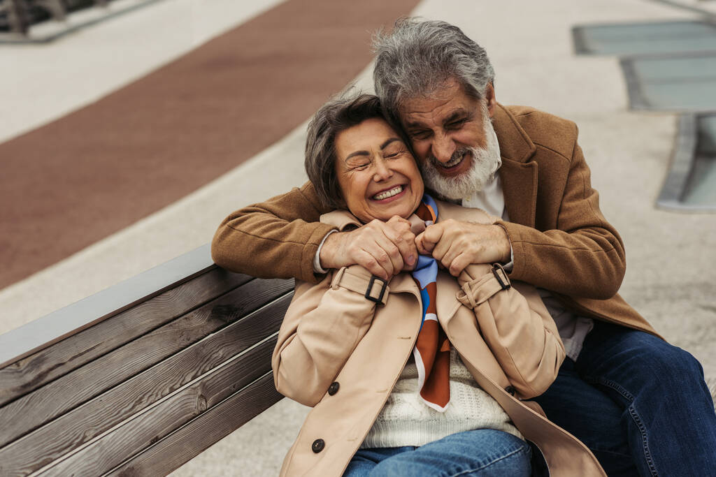 alegre senior hombre en abrigo abrazando anciano esposa sonriendo mientras sentado en banco  - Foto, Imagen