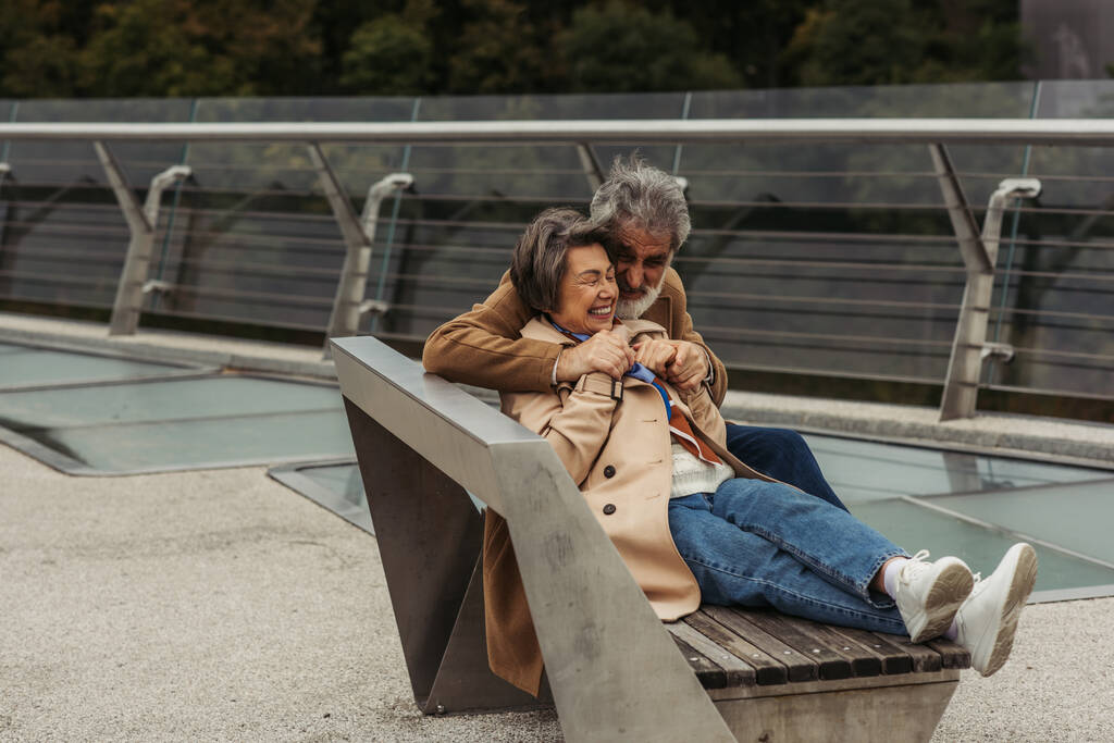 felice uomo anziano abbracciare moglie positiva mentre seduto sulla panchina vicino ponte guardia ferroviaria   - Foto, immagini