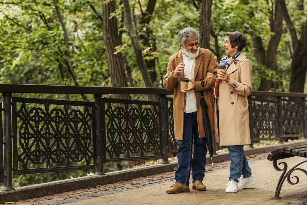 full length of cheerful retired couple in beige coats holding paper cups and walking in park  - Photo, Image