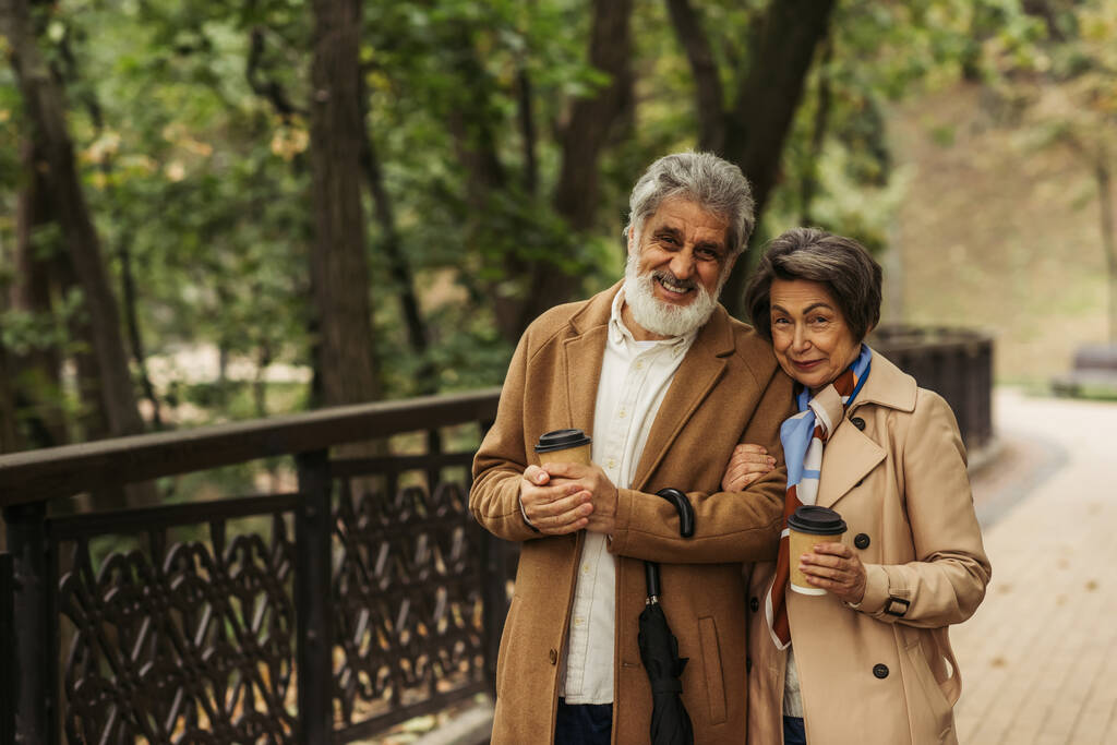 joyeux couple de retraités en manteaux beiges tenant des tasses en papier et marchant dans le parc  - Photo, image
