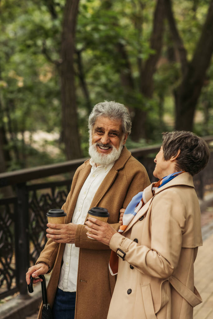 joyful retired couple in beige coats holding paper cups and smiling in park  - Photo, Image