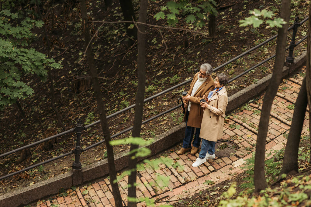 άποψη υψηλής γωνίας χαρούμενα και συνταξιούχος ζευγάρι σε μπεζ παλτά κρατώντας κύπελλα χαρτί, ενώ το περπάτημα στο πάρκο  - Φωτογραφία, εικόνα