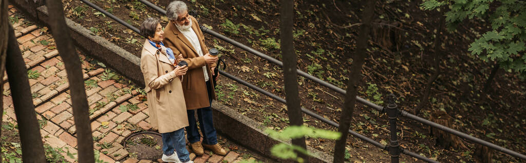 Parkta yürürken ellerinde kağıt bardaklarla gezen neşeli ve emekli çiftin yüksek açılı görüntüsü - Fotoğraf, Görsel