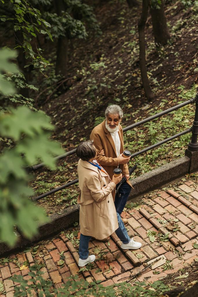 vista de ángulo alto de la pareja mayor complacida en abrigos beige sosteniendo tazas de papel mientras camina en el parque  - Foto, Imagen