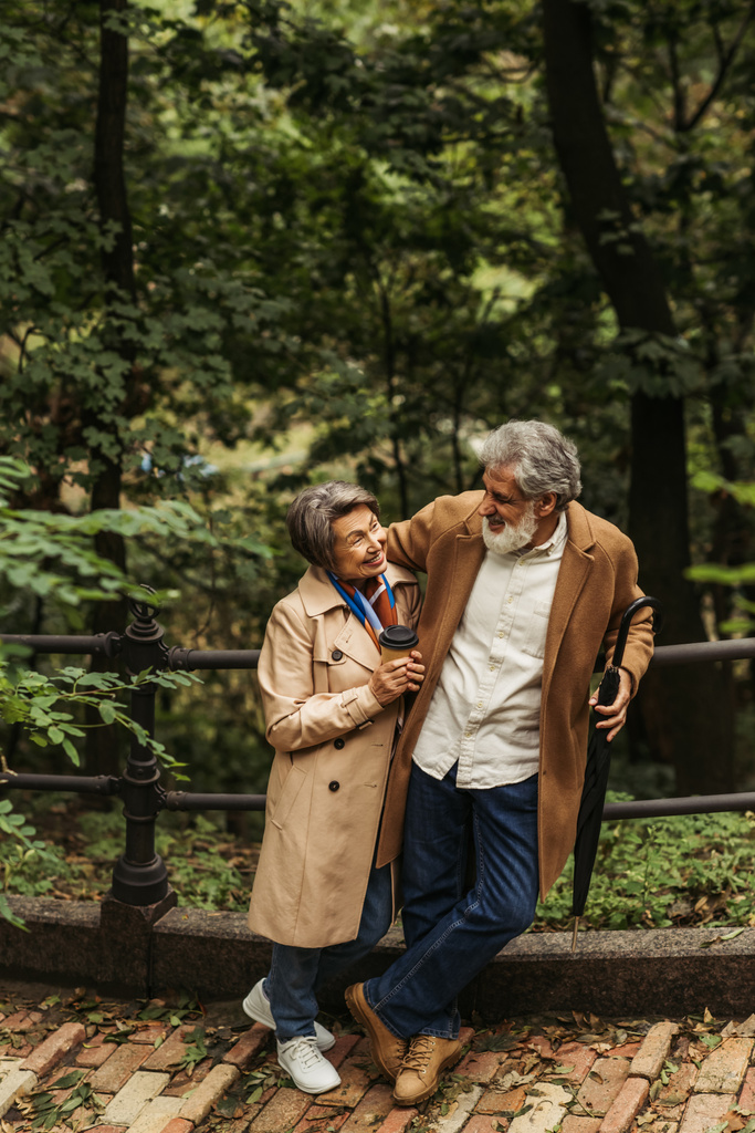 ευτυχισμένος ηλικιωμένος άνδρας κρατώντας ομπρέλα και κοιτάζοντας τη σύζυγο με χάρτινο κύπελλο στο πάρκο - Φωτογραφία, εικόνα