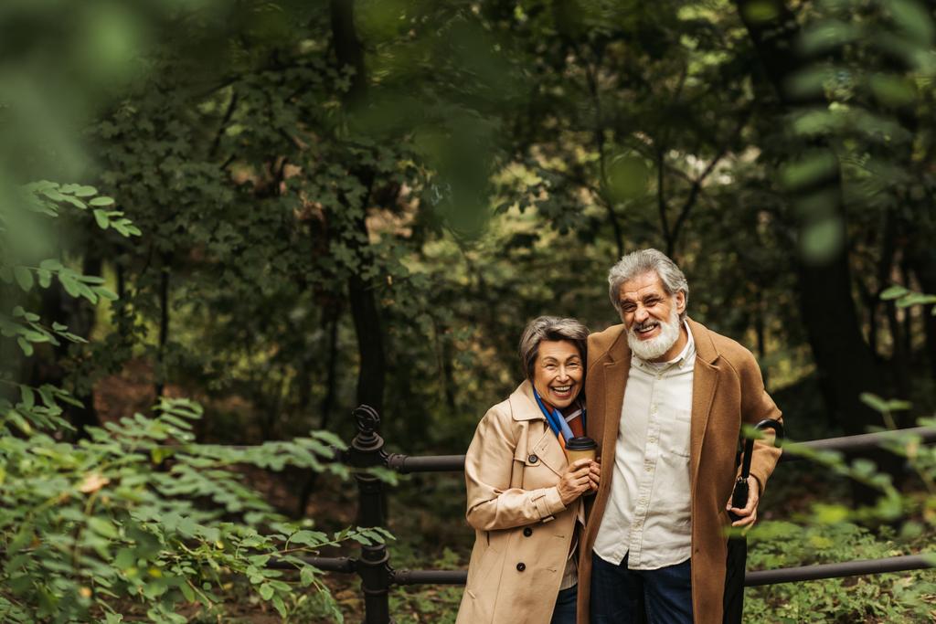 счастливый старший мужчина держит зонтик и стоит рядом с женой с бумажной чашкой в парке - Фото, изображение
