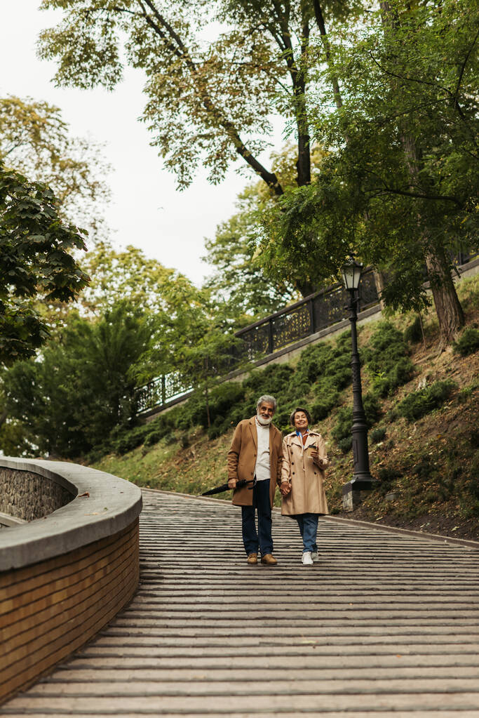 vista completa de la pareja de personas mayores en abrigos caminando con café para ir y paraguas en el parque - Foto, imagen
