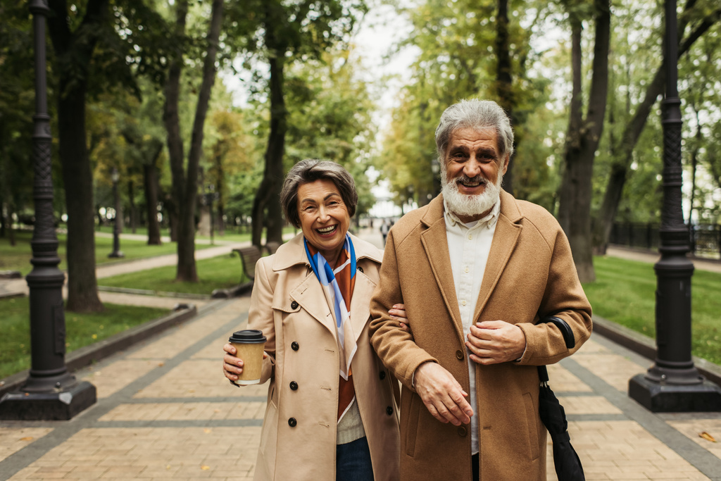 gelukkig senior vrouw holding papier beker terwijl wandelen met stijlvolle man in jas  - Foto, afbeelding