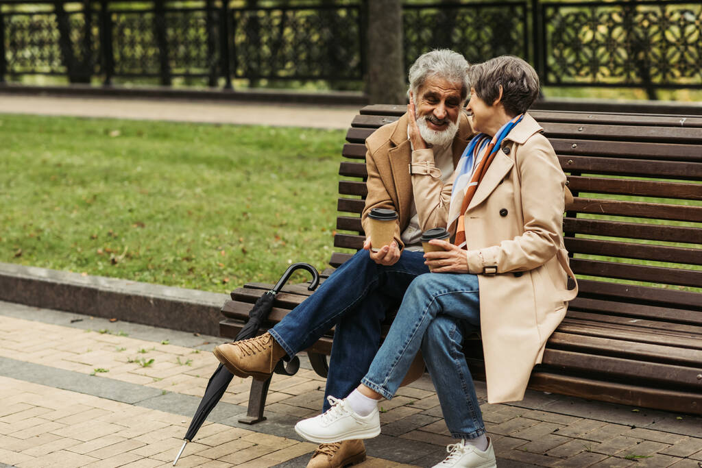 cheerful senior couple in coats holding paper cups and sitting on bench in park  - Photo, Image