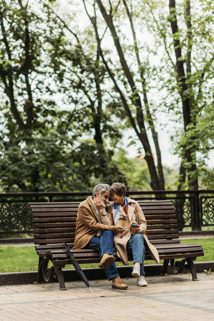 glückliches Rentnerehepaar in Mänteln mit Pappbechern auf Bank im Park sitzend  - Foto, Bild