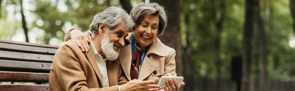 happy senior couple in coats sitting on bench and looking at smartphone, banner - Photo, Image
