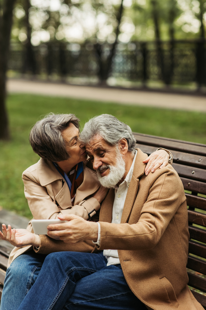 starszy kobieta całuje podczas brodaty mąż w płaszcz biorąc selfie w park - Zdjęcie, obraz