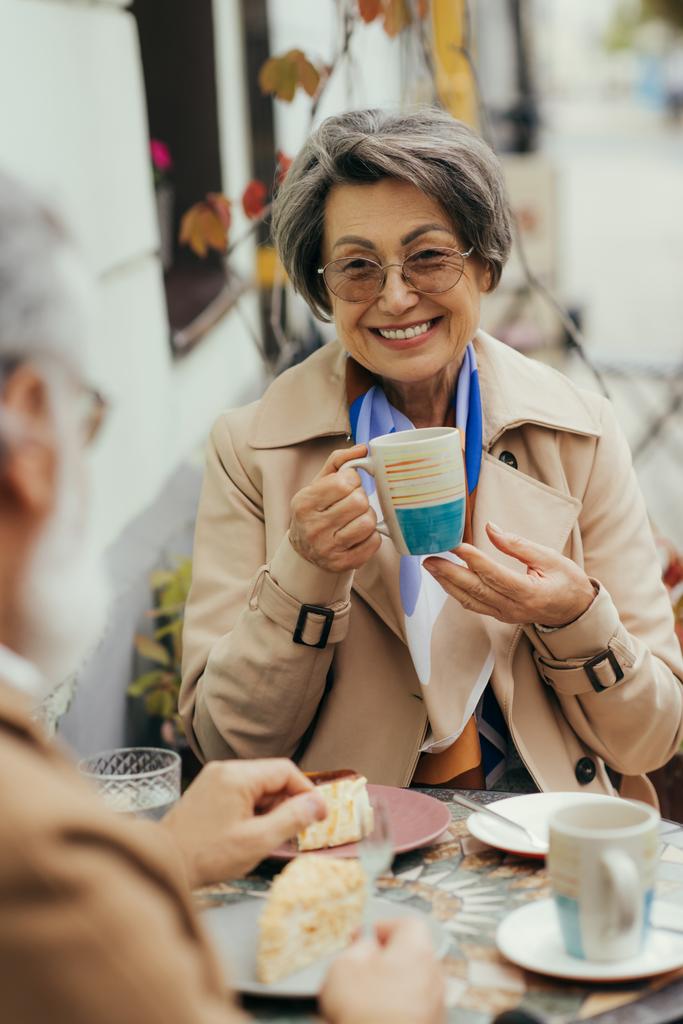 ευτυχισμένη ηλικιωμένη γυναίκα με γυαλιά ηλίου και καμπαρντίνα κρατώντας κύπελλο κατά τη διάρκεια brunch με το σύζυγο στη βεράντα του καφέ - Φωτογραφία, εικόνα